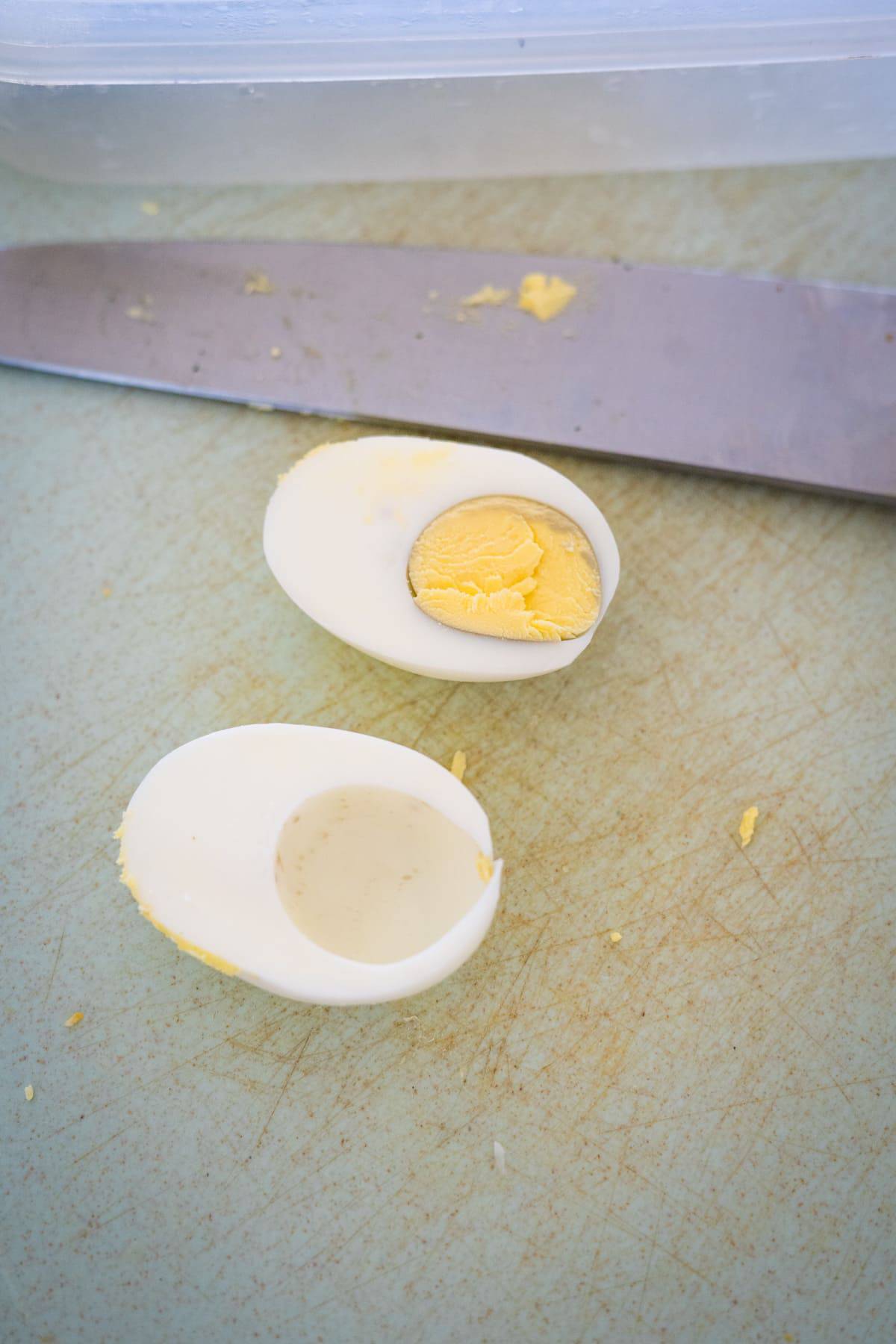 A hard-boiled egg is cut in half on a cutting board, with the yolk removed from one half, ready to be transformed into a delicious keto deviled egg. A knife is placed nearby.