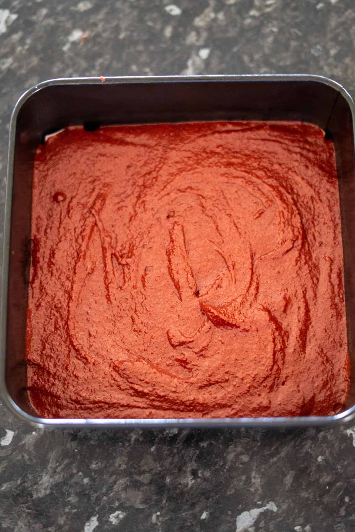 A square metal pan filled with smooth, red-brown batter ready for baking, placed on a dark countertop.