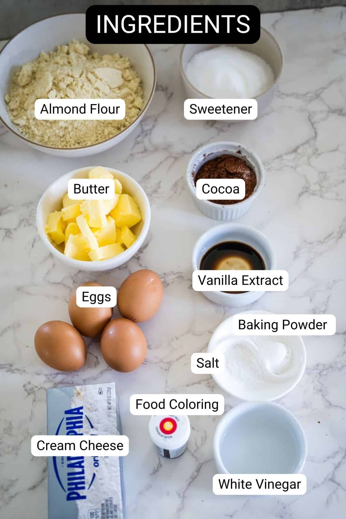 Ingredients on a marble surface: almond flour, sweetener, cocoa, butter, eggs, vanilla extract, baking powder, salt, cream cheese, food coloring, and white vinegar.