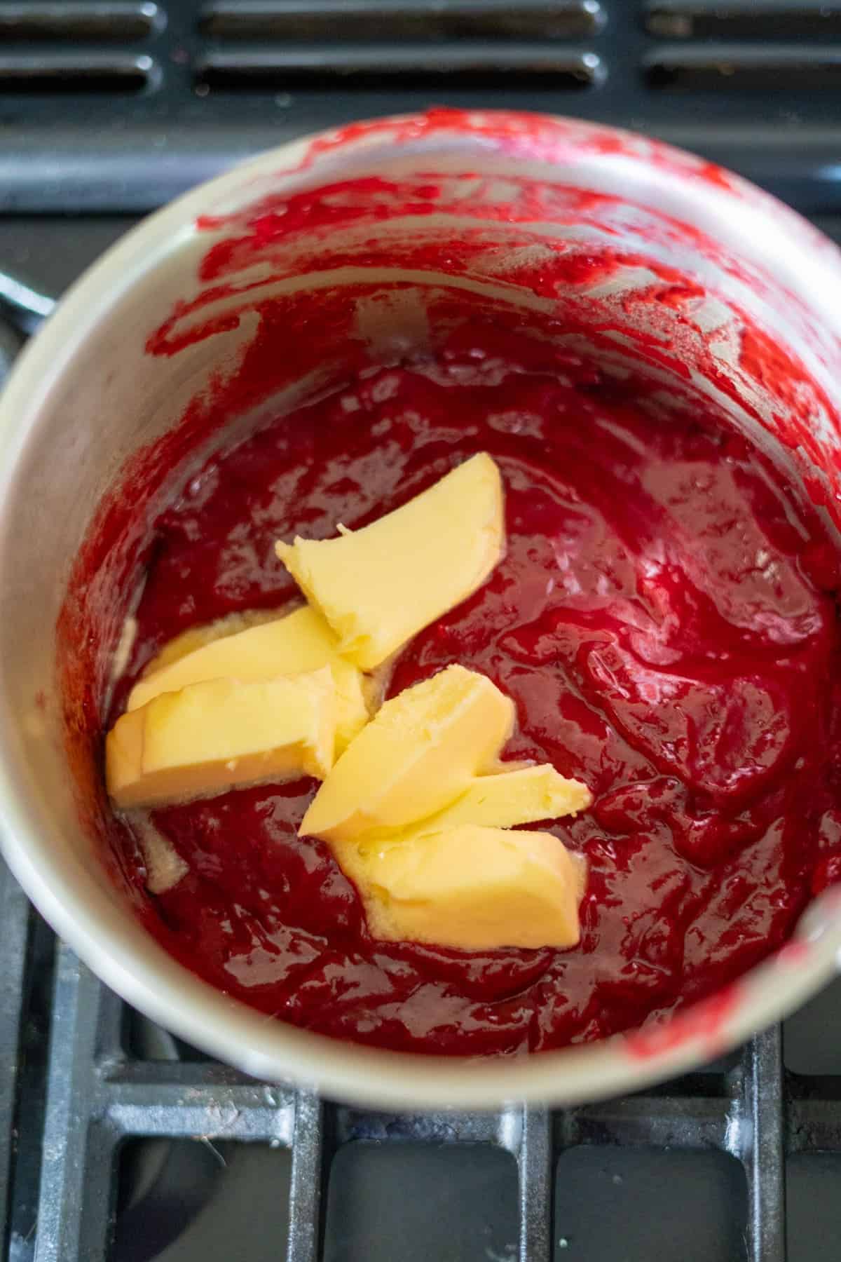 A saucepan containing red berry mixture with chunks of butter on top, placed on a stove.