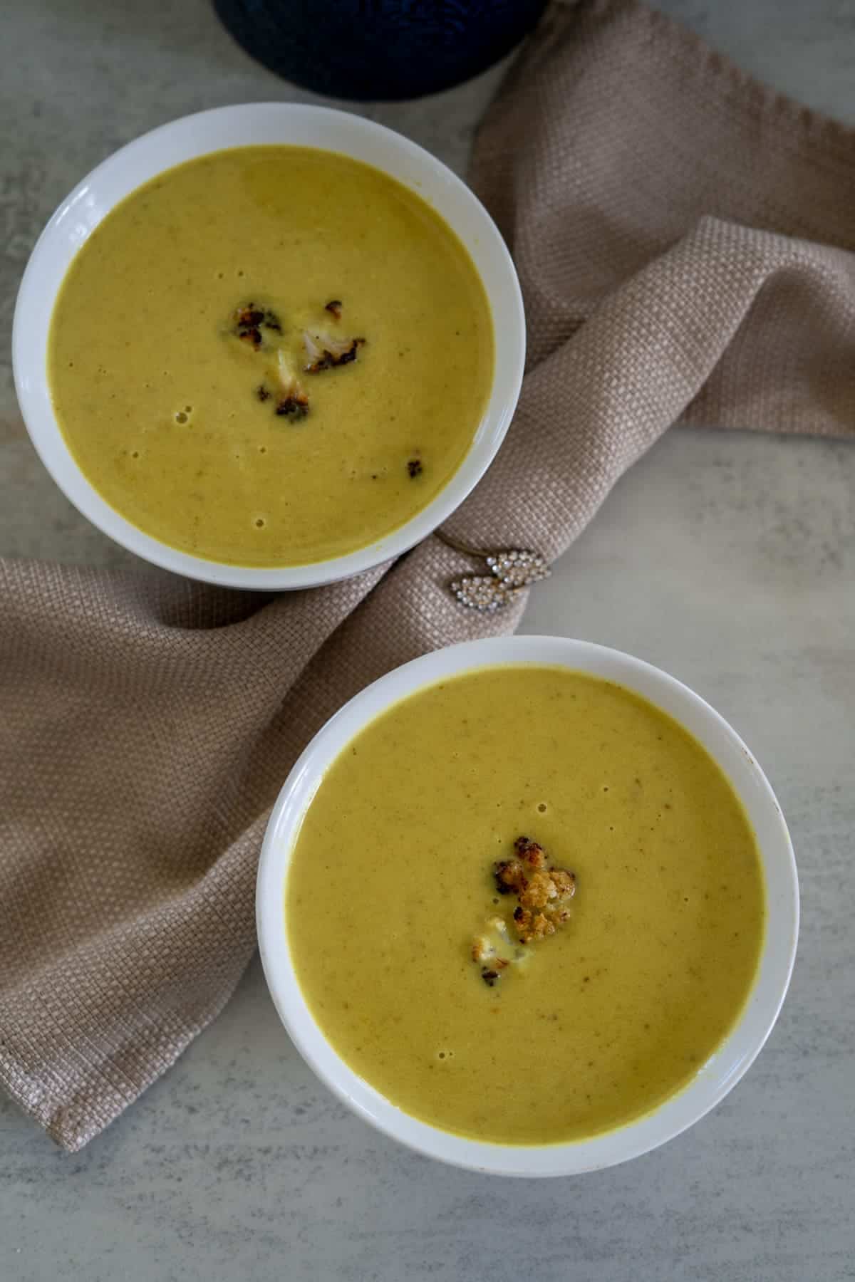 Two white bowls filled with yellow soup, garnished with small pieces of grilled or roasted cauliflower, placed on a folded beige cloth.