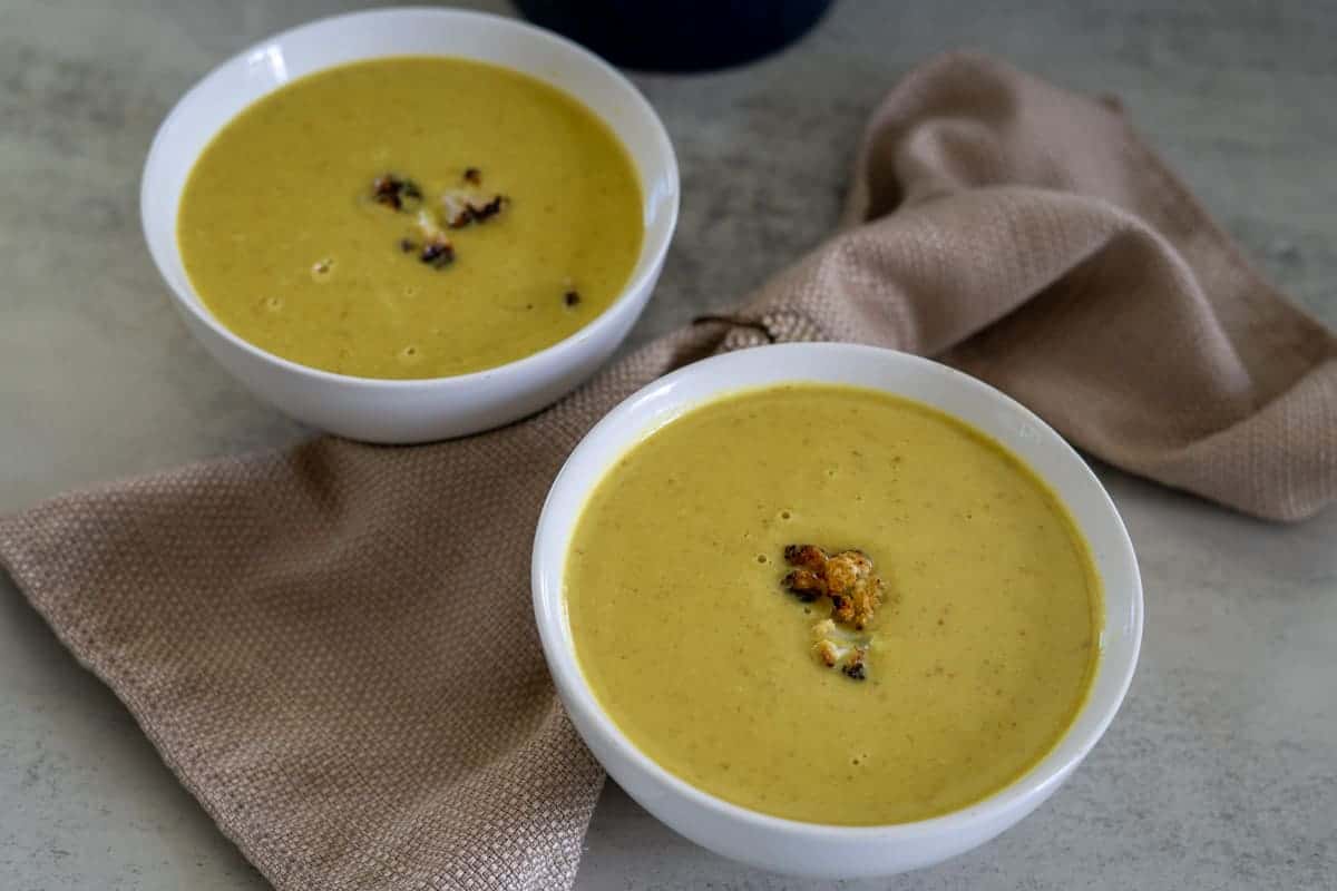 Two white bowls of yellow soup with roasted cauliflower garnish, placed on a gray surface with beige cloth napkins.