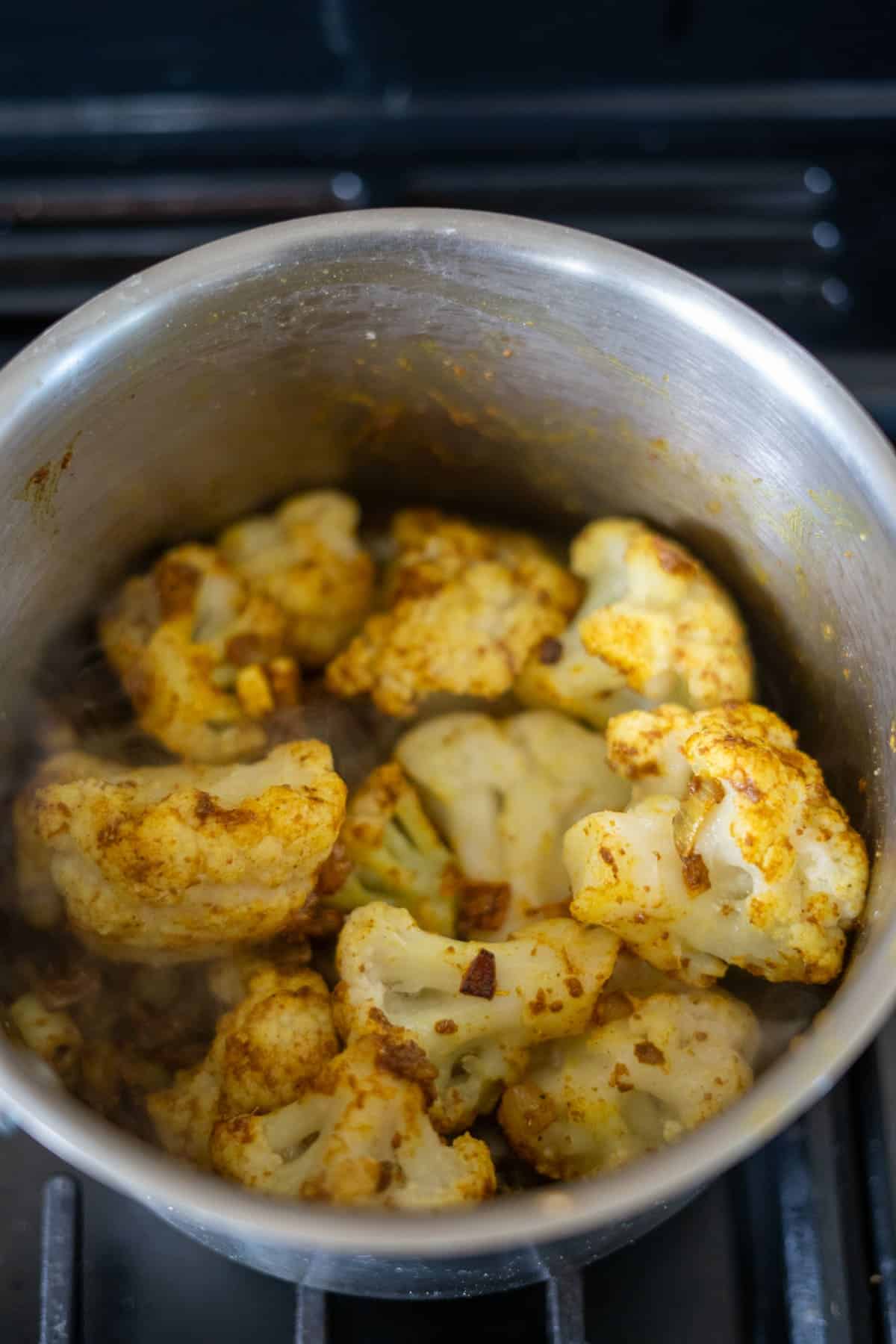 Cauliflower florets seasoned with spices cooking in a stainless steel pot on a stovetop.