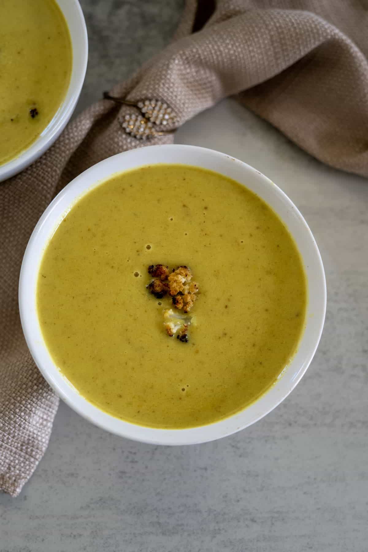 Bowl of creamy green soup garnished with croutons on a light surface, next to a brown cloth.