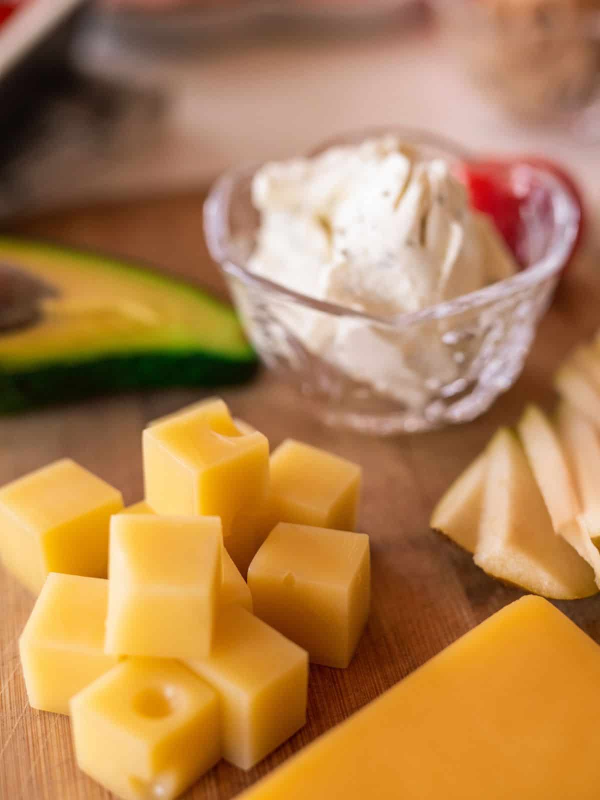 Close-up of cubed cheese, sliced avocado, and a small bowl of creamy cheese on a wooden board—perfect keto travel snacks for your next adventure.