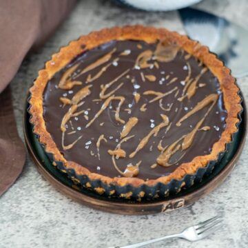 A chocolate tart with a caramel drizzle and sea salt on a wooden plate. A fork and brown cloth are nearby on a textured grey surface.