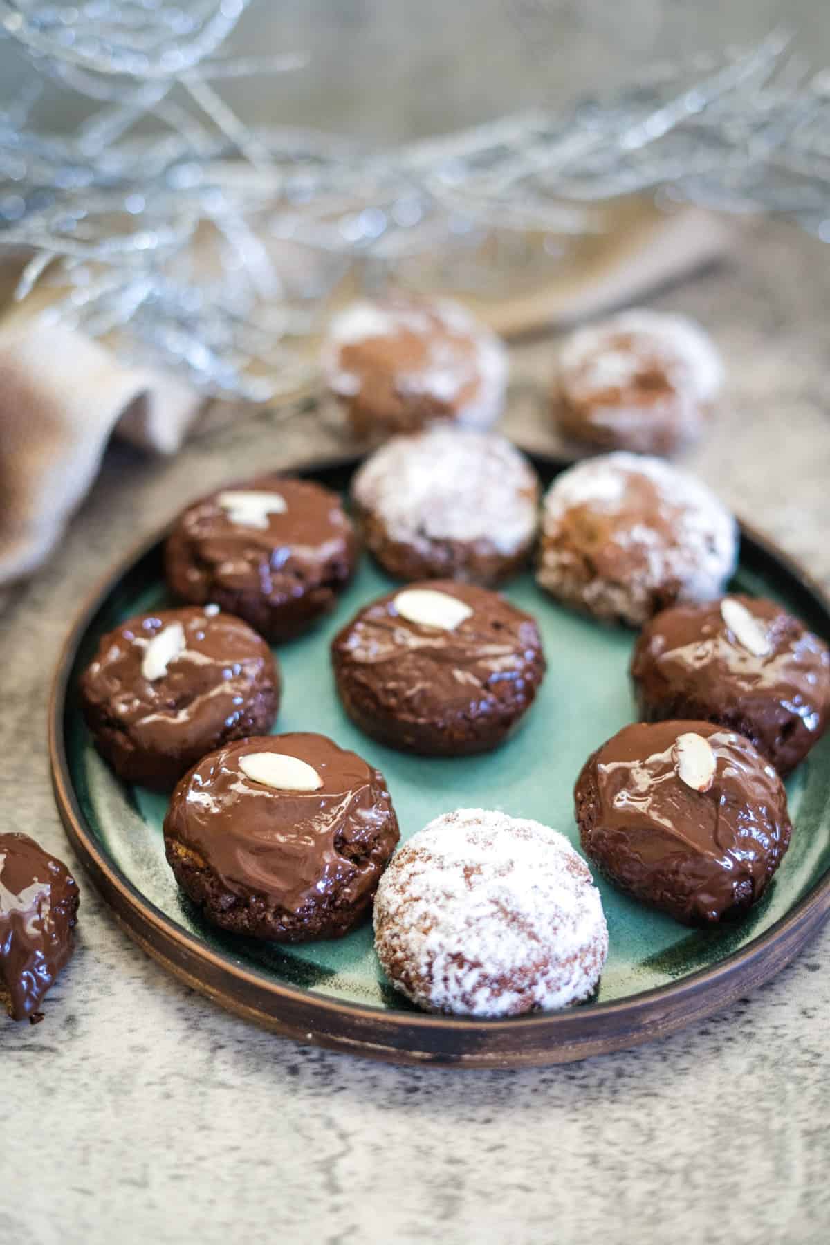 A plate of chocolate-covered cookies, some with almond slices on top and others dusted with powdered sugar, arranged on a green dish.