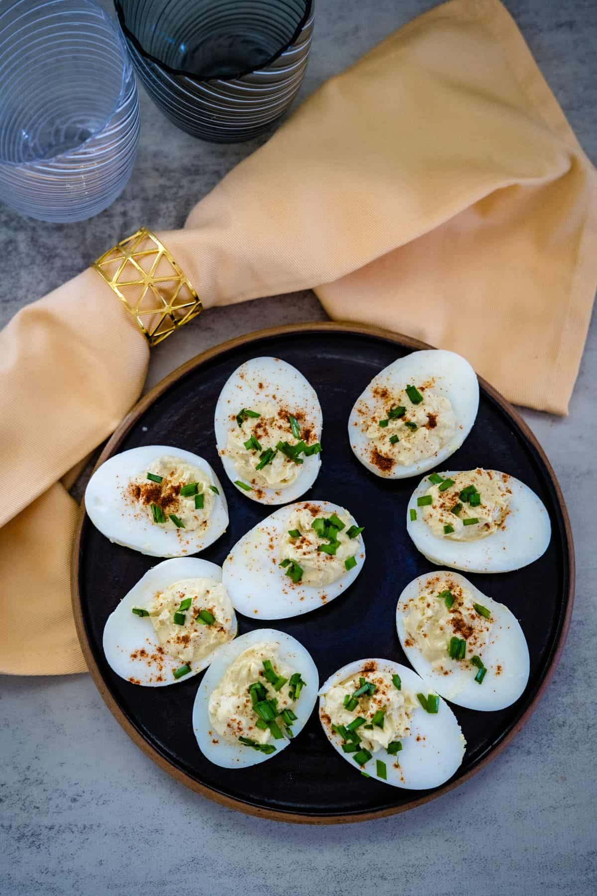 A plate of keto deviled eggs garnished with chives and paprika rests beside a beige napkin with a gold holder and two clear glasses on a gray surface.