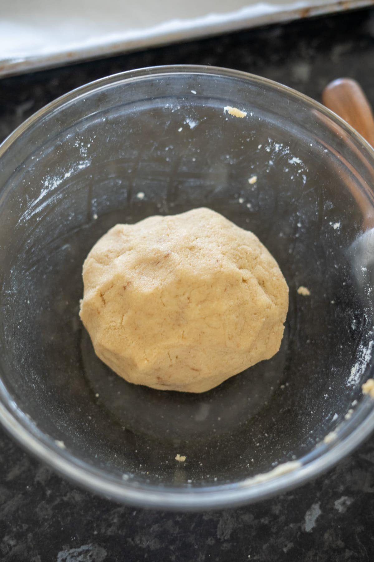 A ball of raw almond flour dough in a clear glass bowl rests on a dark countertop, ready to be transformed into delectable Linzer cookies.