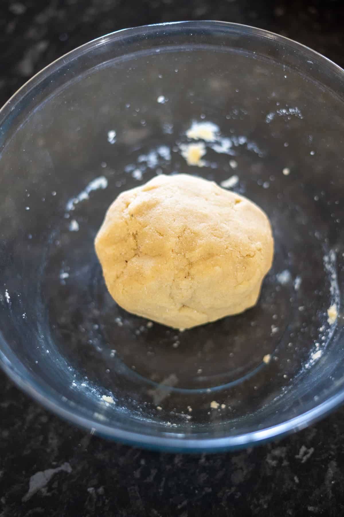 A ball of dough in a glass bowl on a dark countertop.