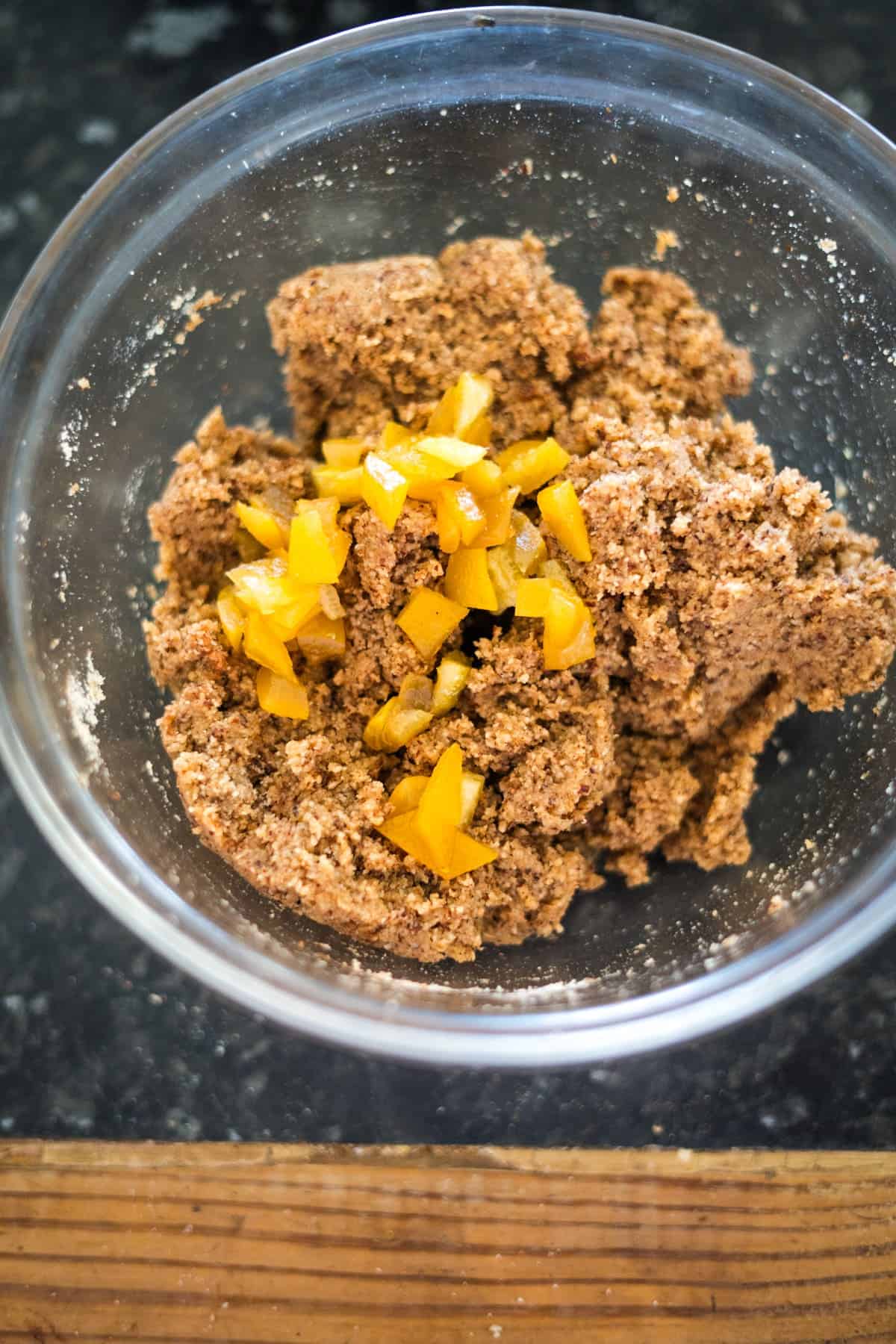 A glass bowl houses a low carb lebkuchen mixture, with hints of yellow chunks atop the brown dough, resting on a dark countertop framed by the wooden board's edge.