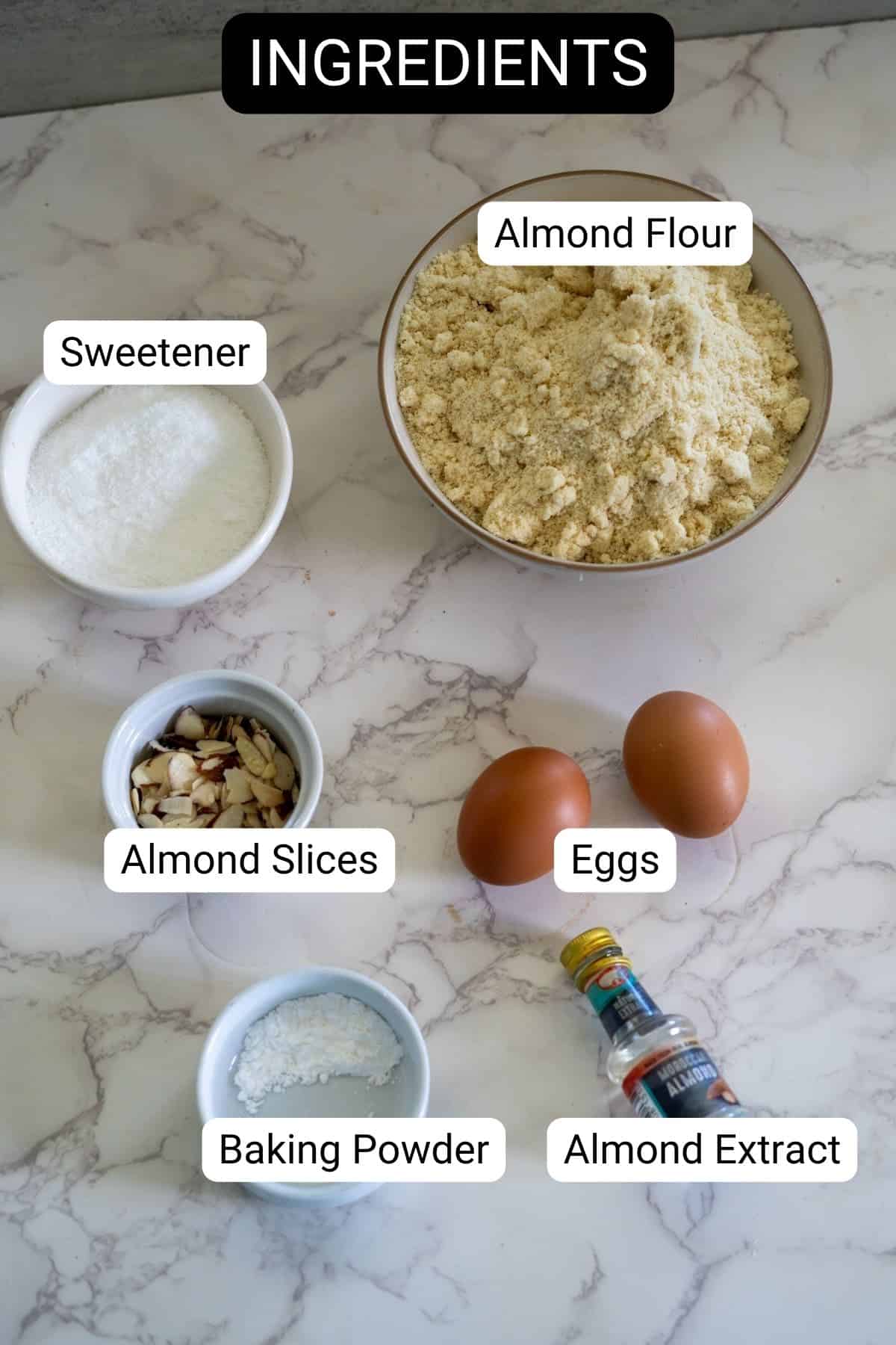 Ingredients for baking: almond flour, eggs, sweetener, almond slices, baking powder, and almond extract on a marble countertop.