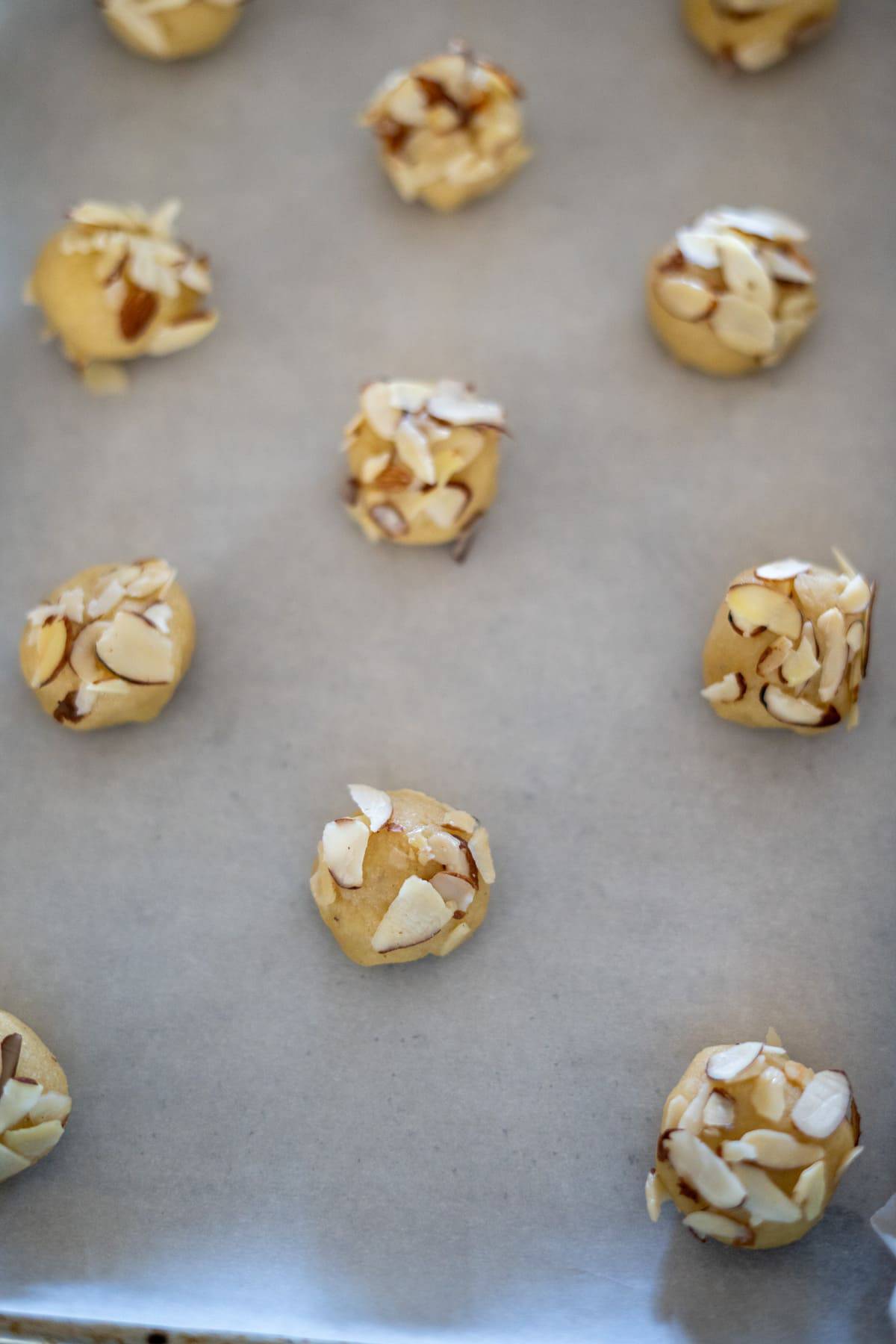 Balls of cookie dough topped with sliced almonds are arranged on a parchment-lined baking sheet.