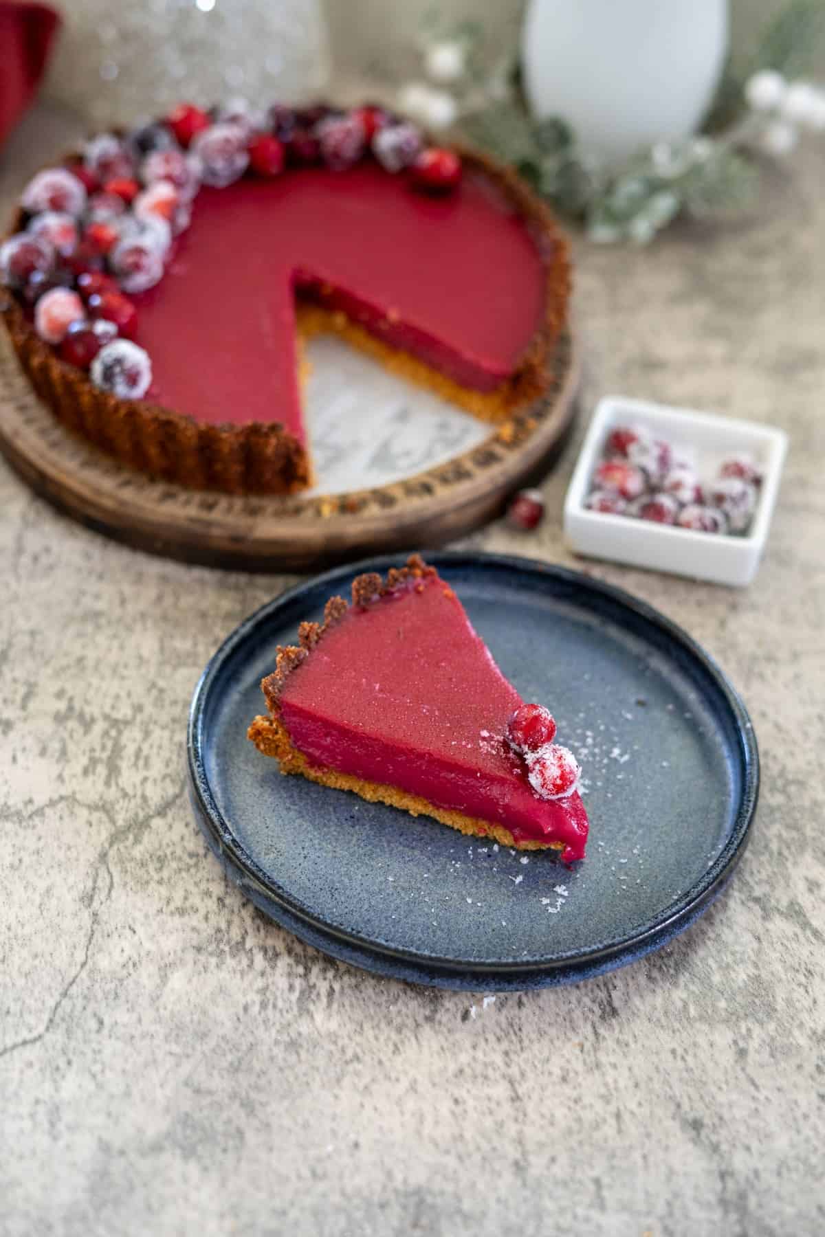 A slice of red tart with cranberries on a blue plate, next to a whole tart on a wooden board and a small dish of sugared cranberries.