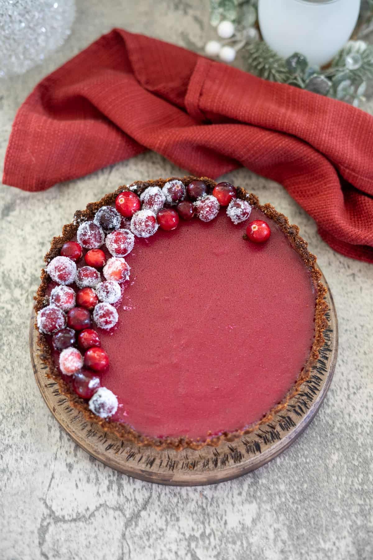 A red cranberry curd tart with almond crust rests on a wooden plate, garnished with sugared cranberries on one side. A red cloth napkin is in the background.