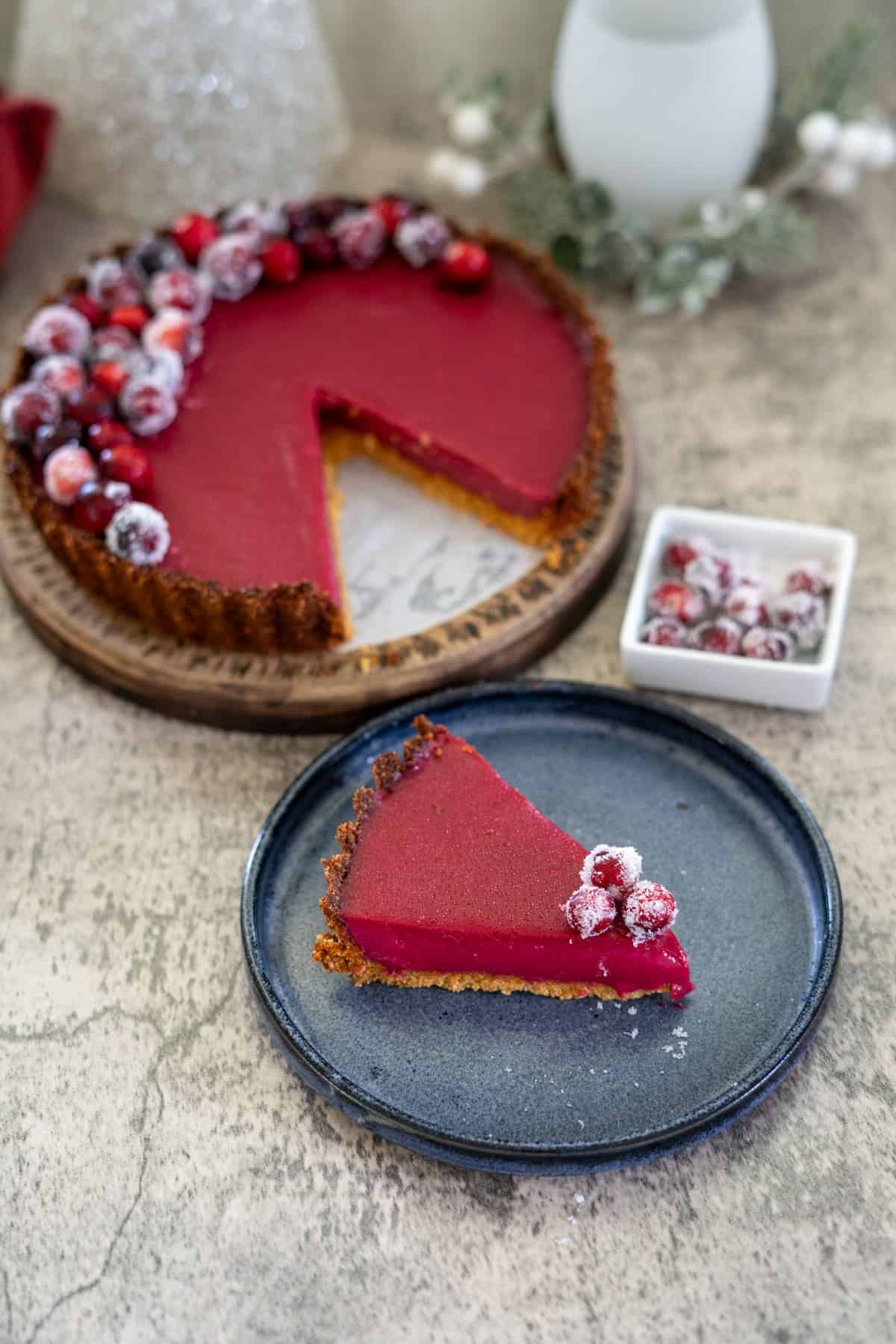 A slice of cranberry curd tart with almond crust sits on a blue plate, while the rest of the tart and sugared cranberries adorn a wooden serving board and fill a small dish in the background.