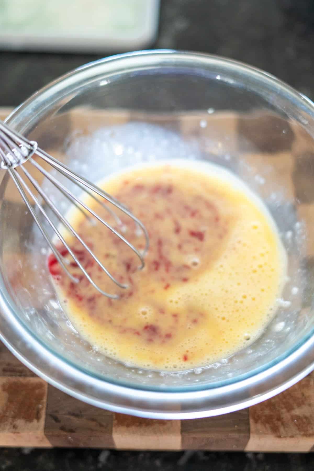 Glass bowl with whisk blending beaten eggs and small pieces of red and brown ingredients on a wooden surface.