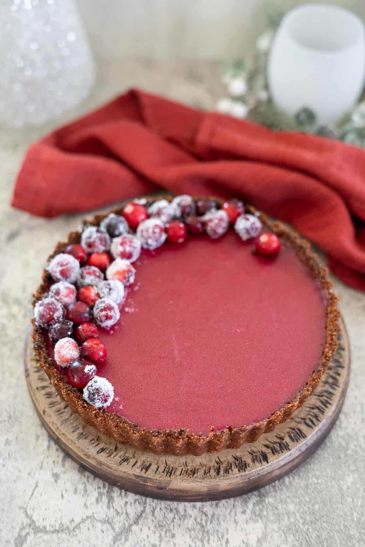 A red tart sits on a wooden plate, topped with frosted cranberries on one side. A red cloth is in the background.