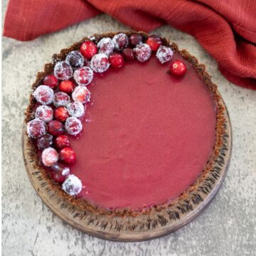 A cranberry tart with a brown crust, topped with sugared cranberries on one side, on a wooden plate beside a red cloth.