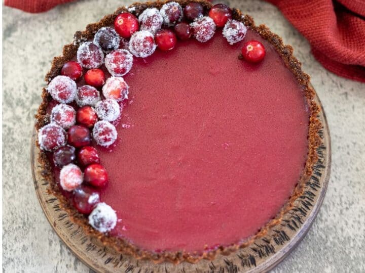 A cranberry tart with a brown crust, topped with sugared cranberries on one side, on a wooden plate beside a red cloth.