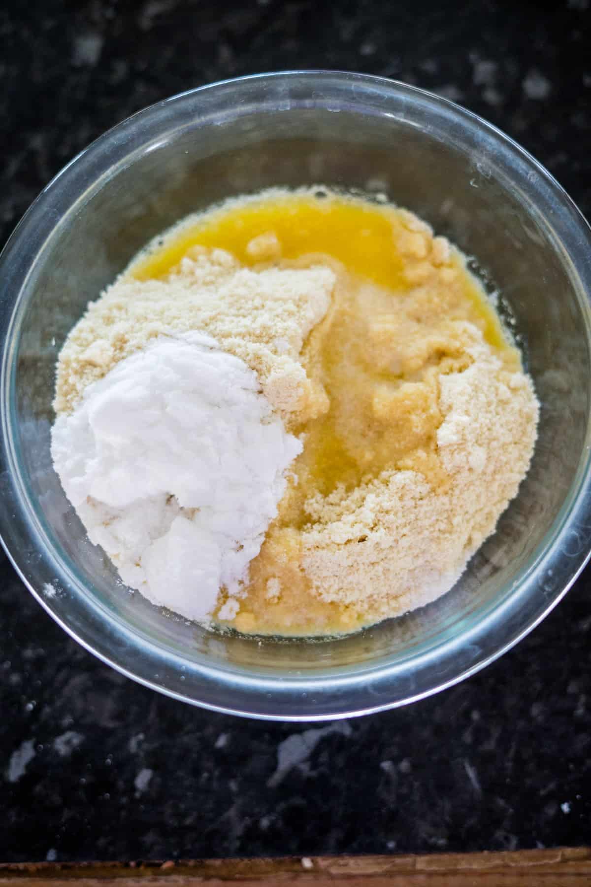 A glass bowl contains almond flour, melted butter, and a scoop of baking powder, placed on a dark countertop.