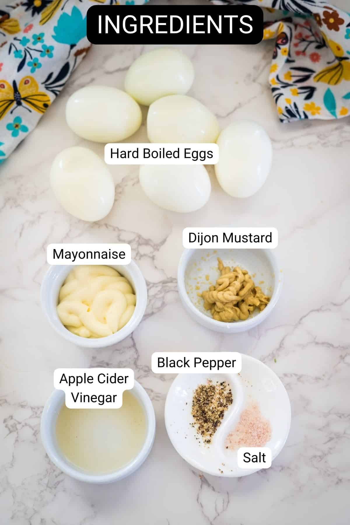 Labeled ingredients for a recipe: hard boiled eggs, mayonnaise, Dijon mustard, apple cider vinegar, black pepper, and salt placed on a marble surface.