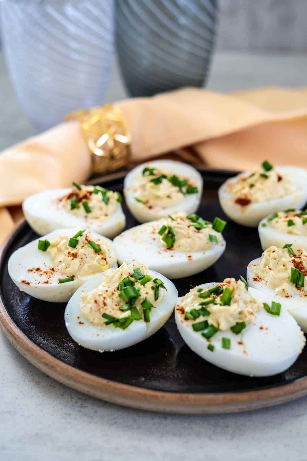 Plate of keto deviled eggs garnished with chopped chives and paprika, arranged on a dark round plate. Two glass cups and a folded napkin lie elegantly in the background.