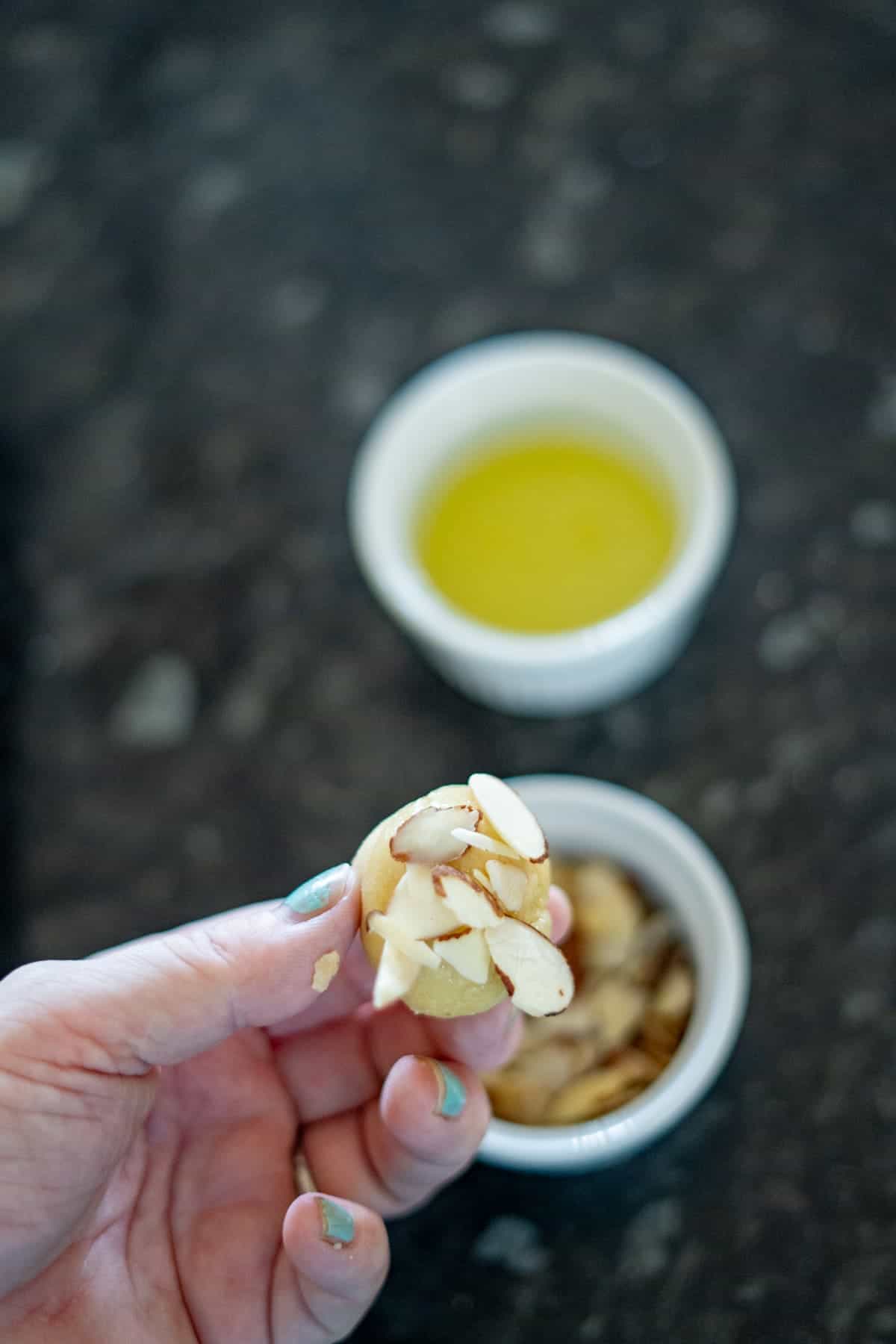 Hand holding a small cookie topped with sliced almonds, with a bowl of almonds and a cup of yellow liquid in the background on a dark surface.