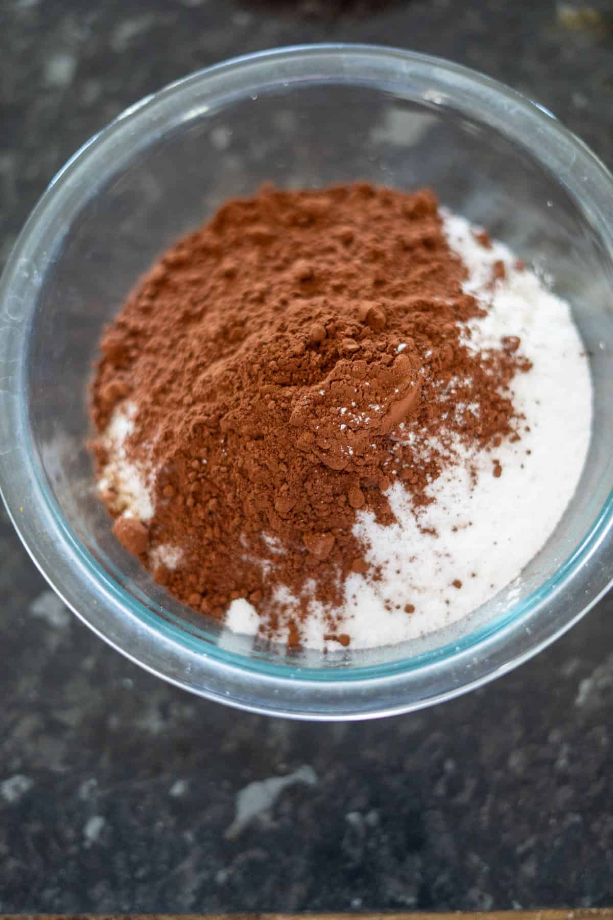 Glass bowl with white flour and brown cocoa powder on a dark countertop.