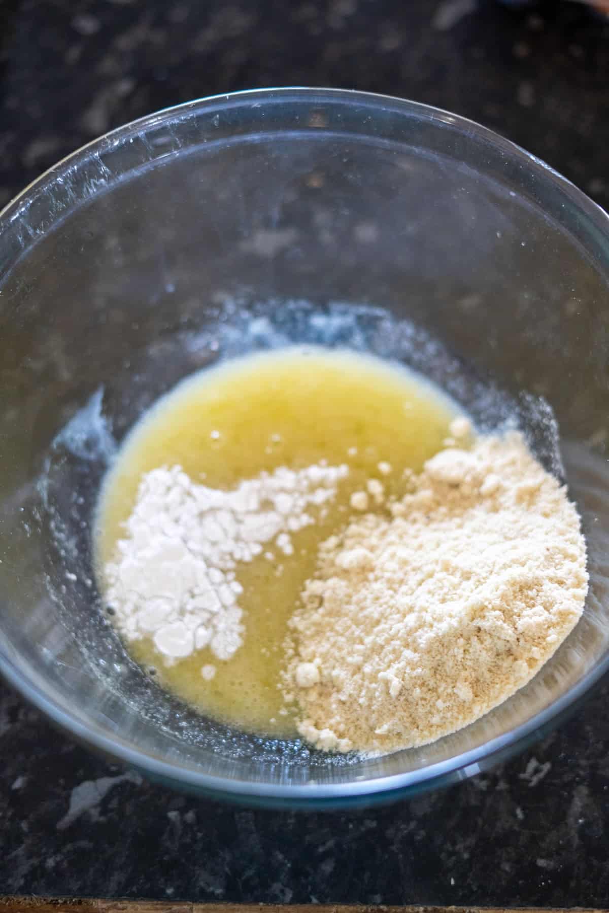 A glass bowl with melted butter, flour, and almond flour on a dark countertop.