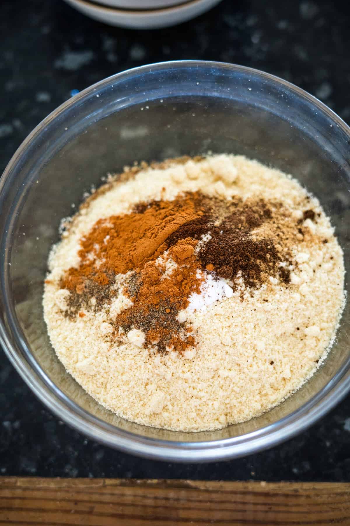 A clear glass bowl contains a mixture of almond flour, cinnamon, nutmeg, and other powdered ingredients perfect for crafting low carb lebkuchen on a dark countertop.