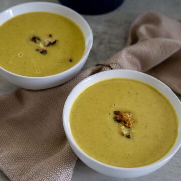 Two bowls of creamy yellow soup garnished with roasted cauliflower, placed on a textured beige napkin on a neutral surface.