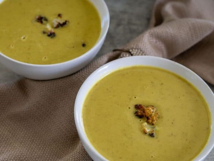 Two bowls of creamy yellow soup garnished with roasted cauliflower, placed on a textured beige napkin on a neutral surface.