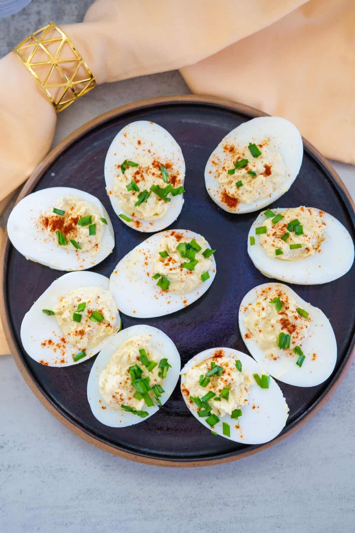 A plate of deviled eggs garnished with chives and paprika, set on a table with a beige napkin and gold napkin ring.