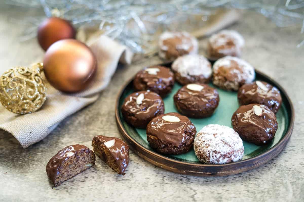 A plate of chocolate-covered cookies, reminiscent of low carb lebkuchen, some dusted with powdered sugar, sits next to decorative ornaments and a burlap cloth on a gray surface.