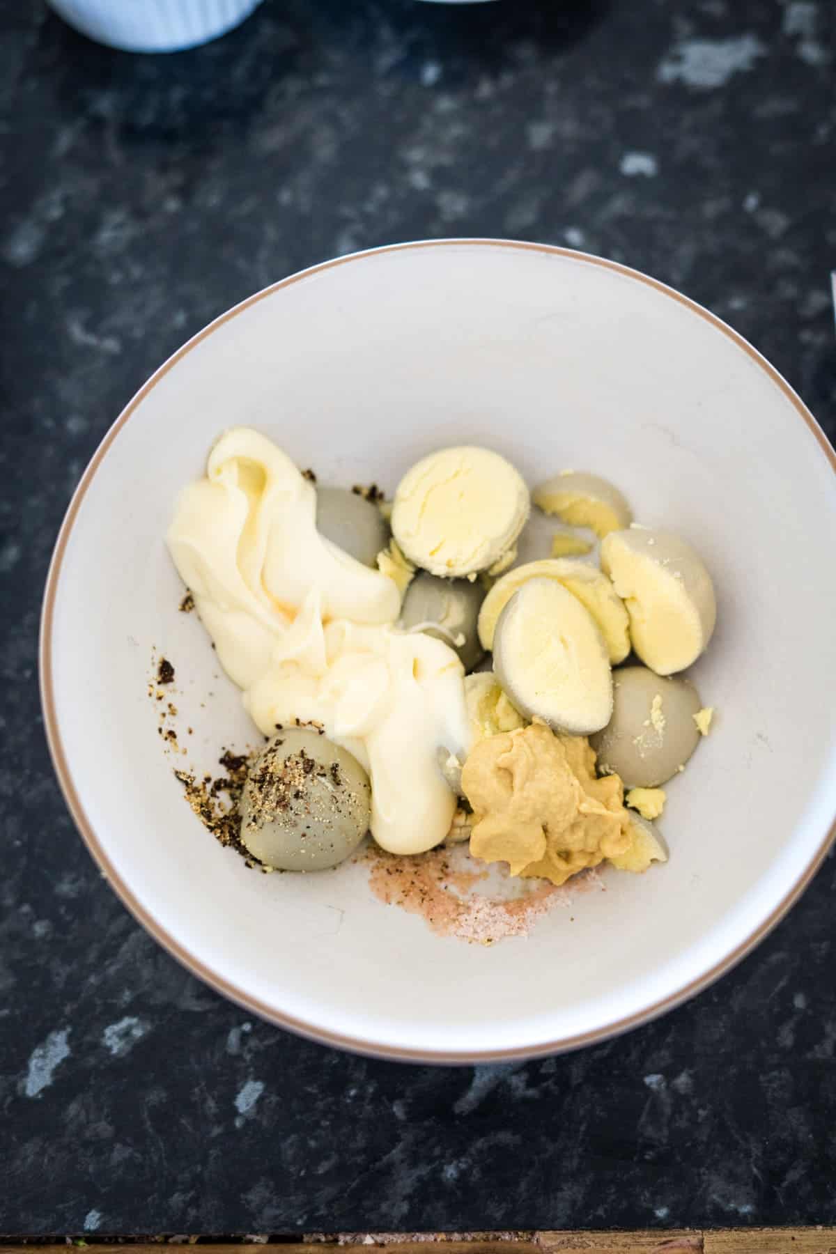 A bowl containing scoops of butter, a dollop of mayonnaise, mustard, pepper, and a pinch of salt sits on a dark countertop, ready to be mixed into the perfect base for keto deviled eggs.