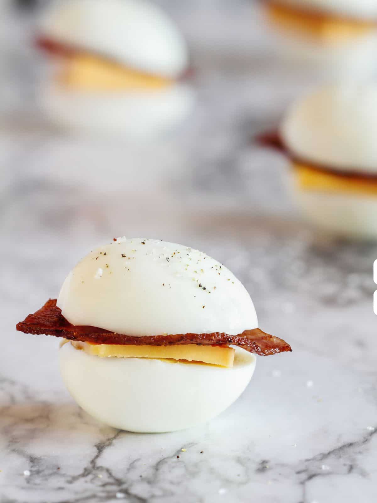 Close-up of a hard-boiled egg slider with bacon and cheese on a marble surface, perfect as keto travel snacks. Similar sliders are blurred in the background, offering a delectable glimpse into your next on-the-go meal.