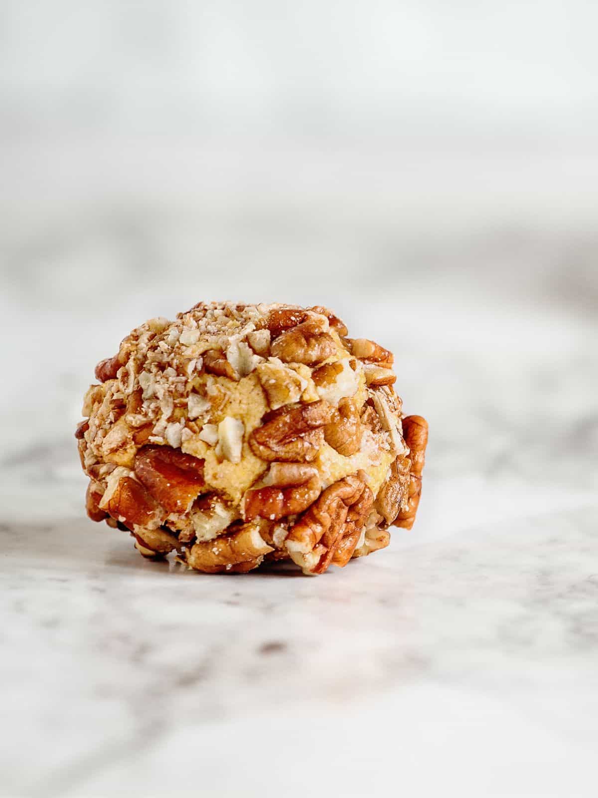 A cheese ball, perfect for keto travel snacks, is coated with chopped nuts and rests elegantly on a marble surface.