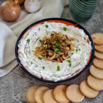 A bowl of onion dip garnished with caramelized onions and chives, surrounded by round crackers. Onions, garlic, and a cloth napkin are in the background alongside two green glasses.