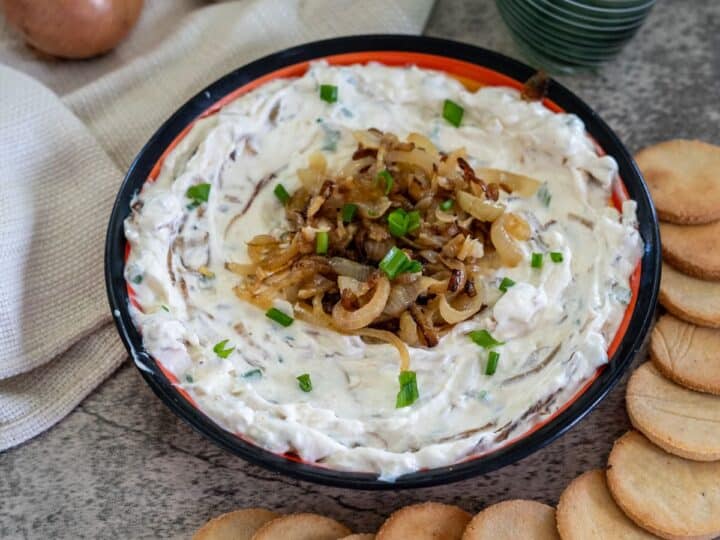 A bowl of onion dip garnished with caramelized onions and chives, surrounded by round crackers. Onions, garlic, and a cloth napkin are in the background alongside two green glasses.