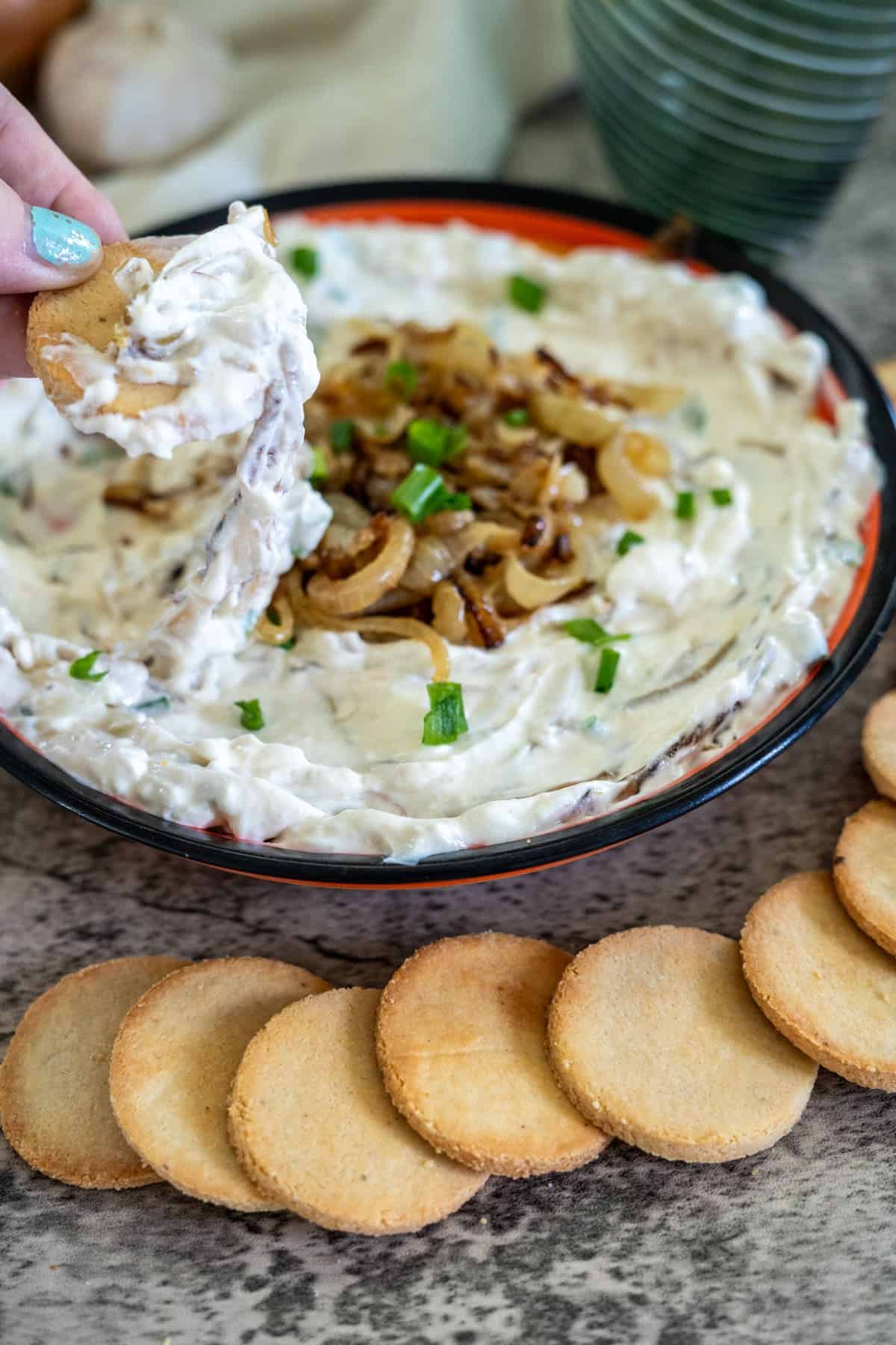 A hand dipping a cracker into a bowl of creamy onion dip, garnished with caramelized onions and chopped green onions. Crackers are arranged in front of the bowl.