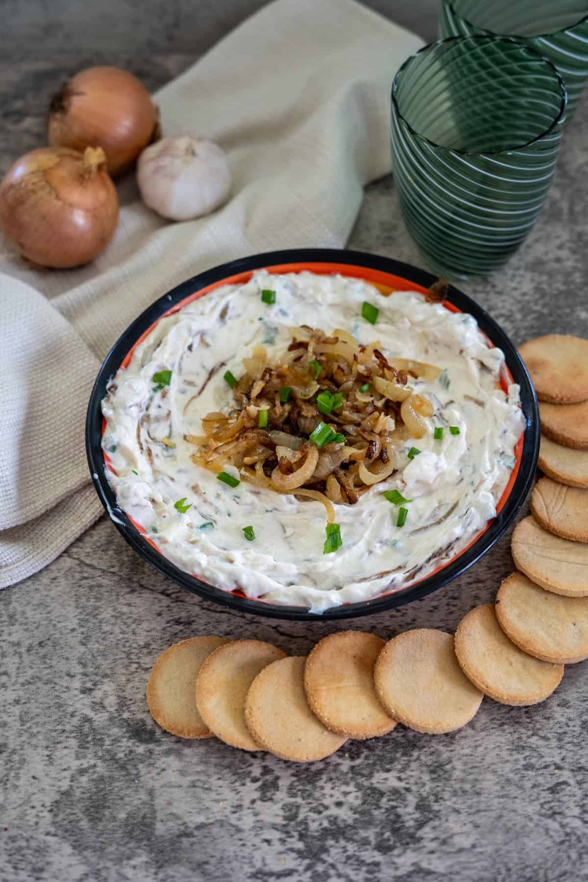 A bowl of onion dip garnished with caramelized onions and chives, surrounded by round crackers. Onions, garlic, and a cloth napkin are in the background alongside two green glasses.