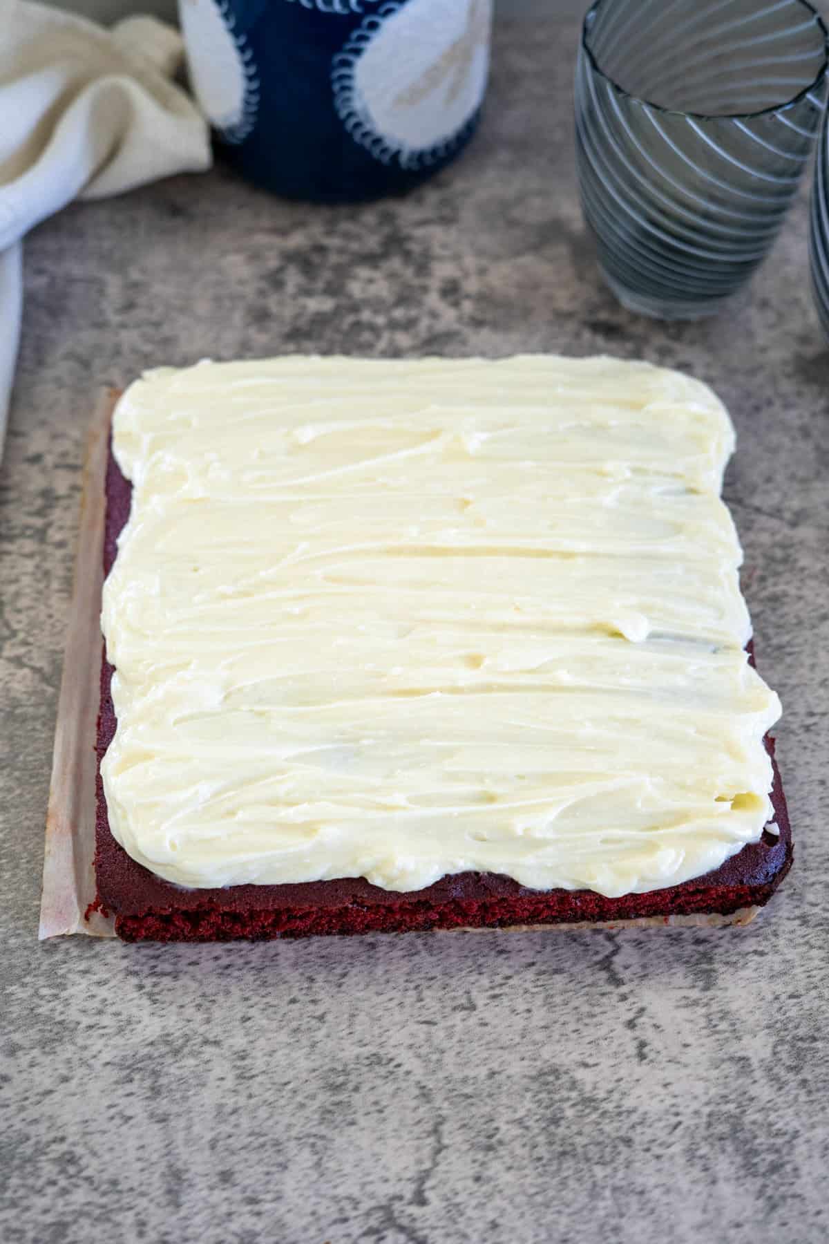 A square red velvet cake on a gray surface, topped with a thick layer of cream cheese frosting.