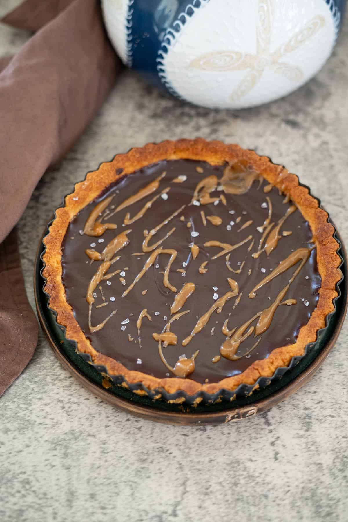 Keto chocolate tart with a glossy ganache topping, drizzle of caramel, and a sprinkle of sea salt on a gray surface near a brown cloth and a decorative ceramic item.