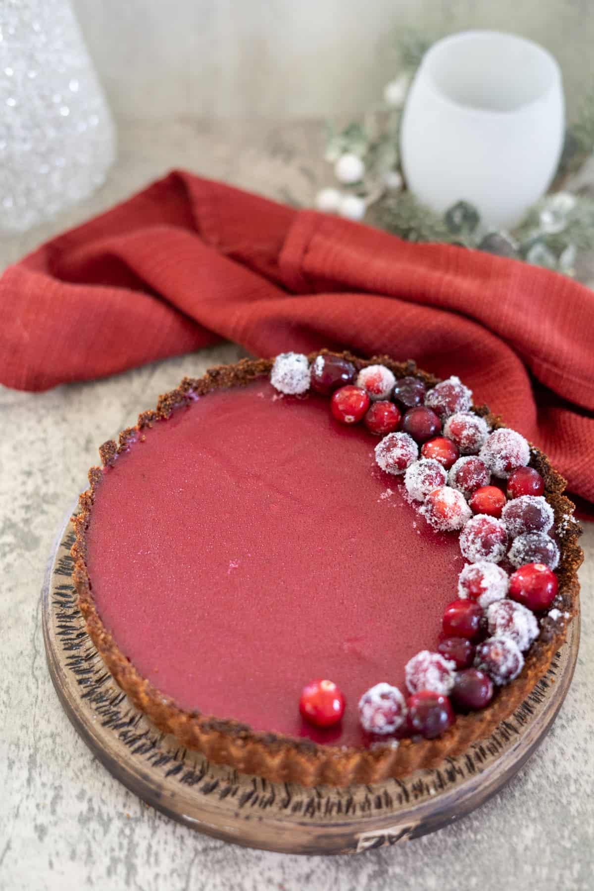 A cranberry curd tart with almond crust sits elegantly on a wooden plate, adorned with fresh cranberries and a dusting of powdered sugar. A vibrant red cloth and pristine white glass complete the picturesque setting in the background.