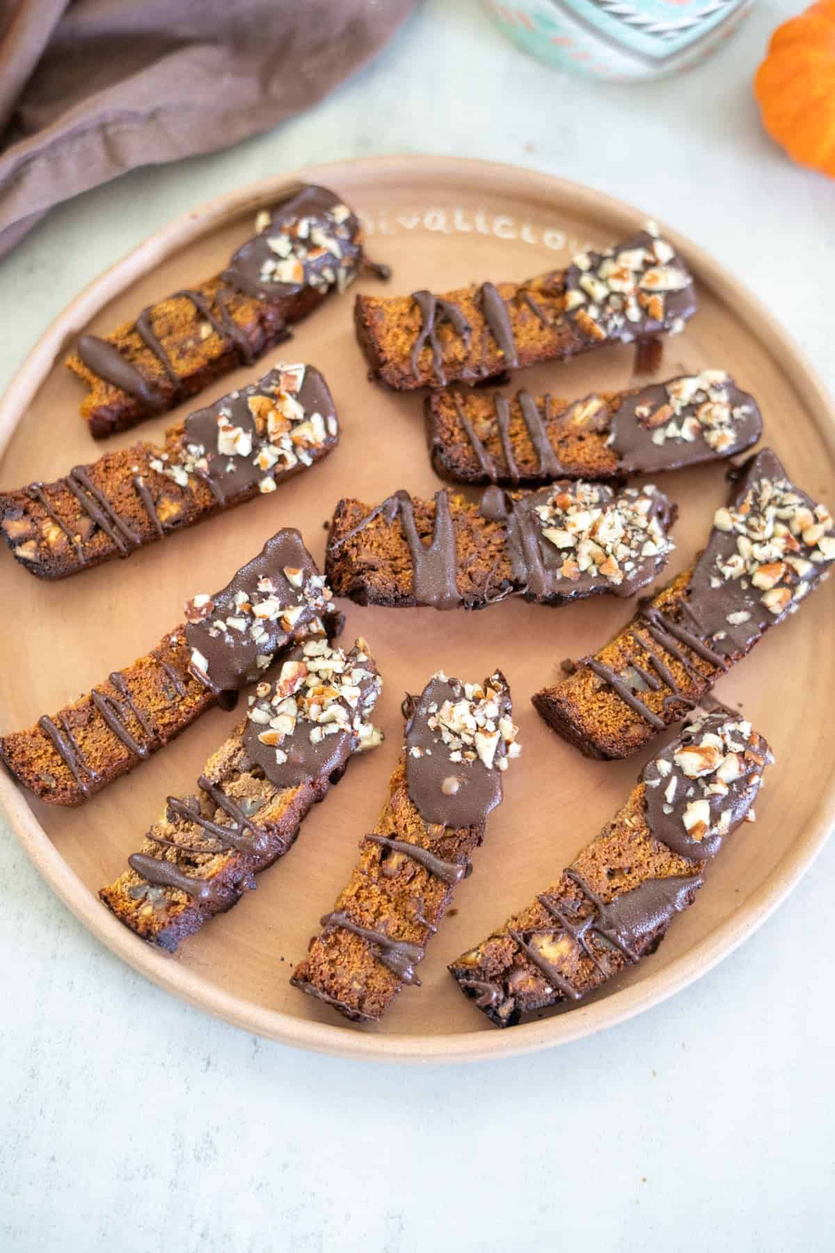 A plate of pumpkin biscotti, each piece decadently chocolate-dipped and sprinkled with chopped nuts, rests invitingly on a light surface.