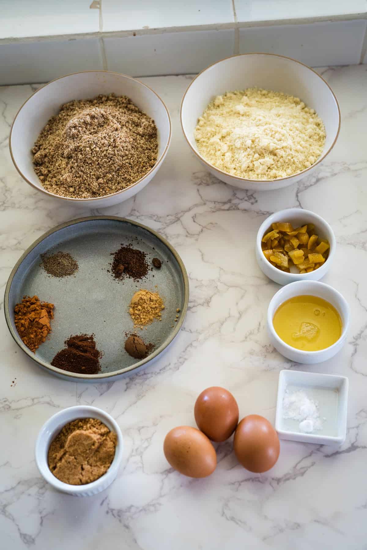 Various baking ingredients are arranged on a marble surface, featuring bowls of low carb flour, spices on a plate reminiscent of lebkuchen, eggs, and small dishes of diced fruits, liquid, and baking powder.