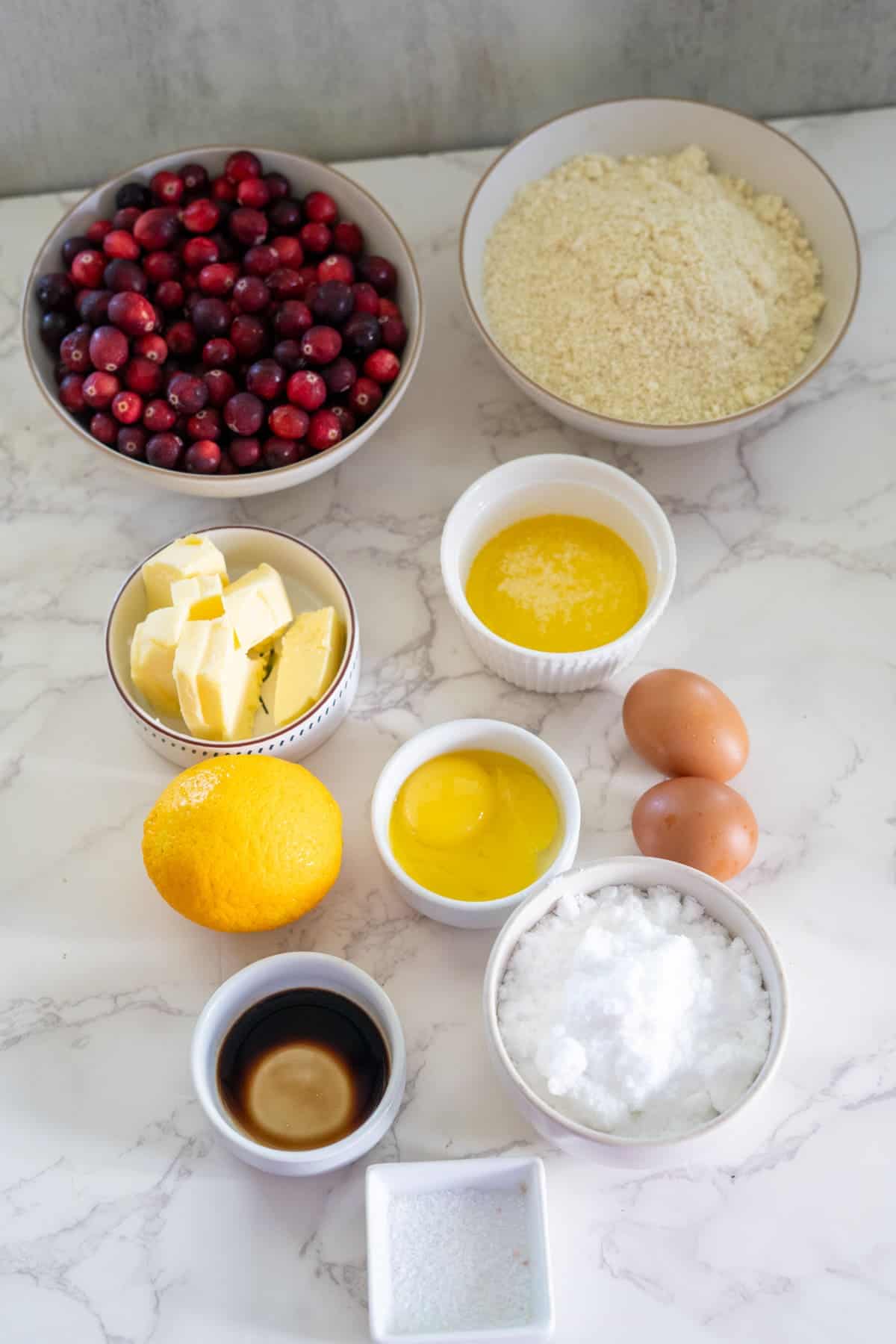 A marble countertop displays bowls containing cranberries, flour, butter, sugar, eggs, melted butter, an orange, vanilla extract, and salt.