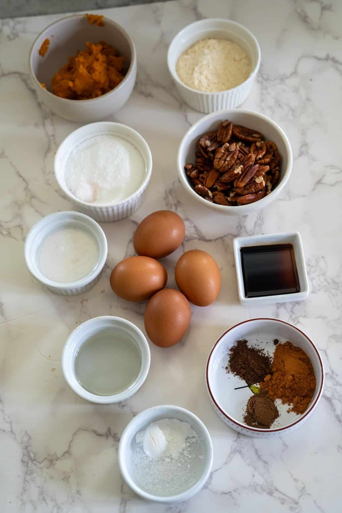 Ingredients for a pumpkin pecan recipe on a marble surface: eggs, pecans, flour, sugar, pumpkin puree, vanilla extract, spices, baking soda, and salt.