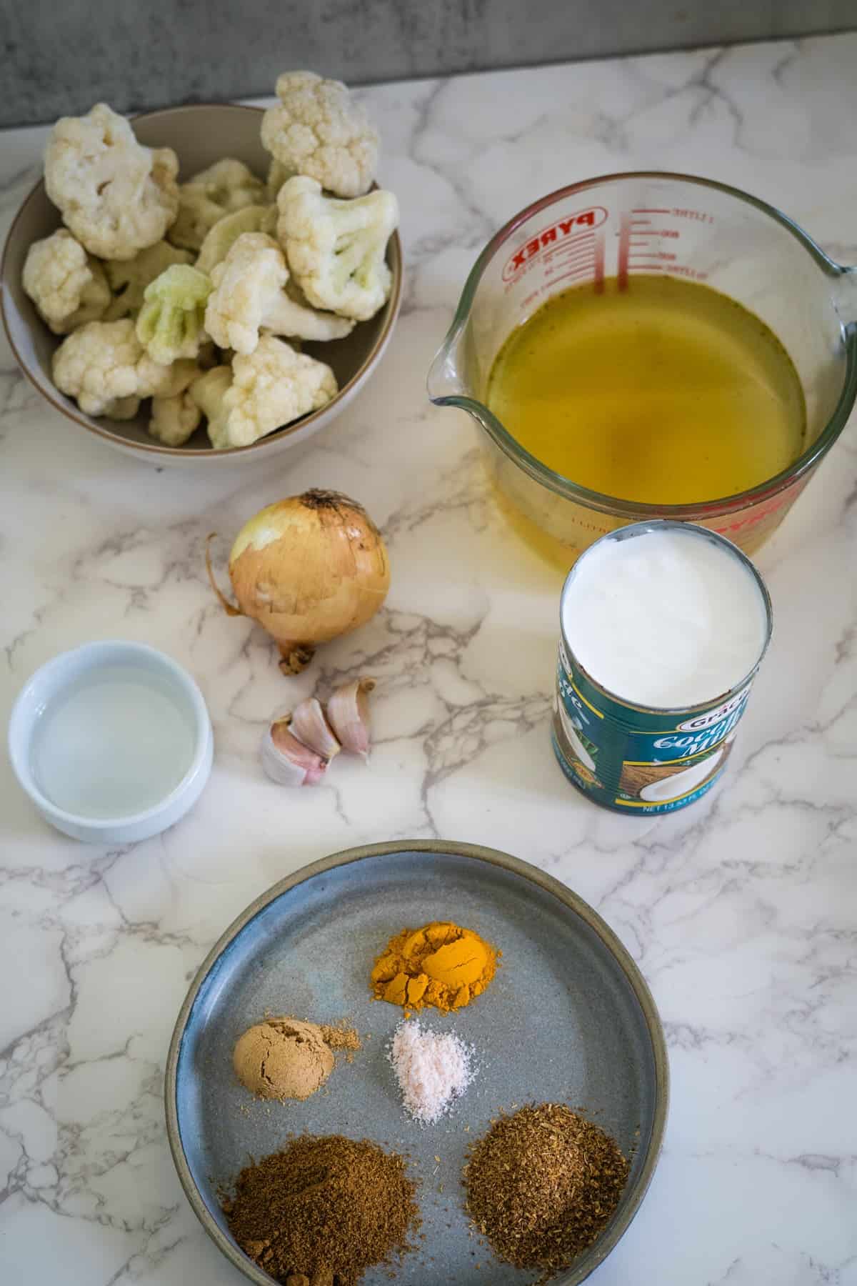 Ingredients for cooking: cauliflower, onion, garlic, broth in a measuring cup, coconut milk, a small bowl of water, and a plate with spices: turmeric, salt, cumin, coriander, and curry powder.